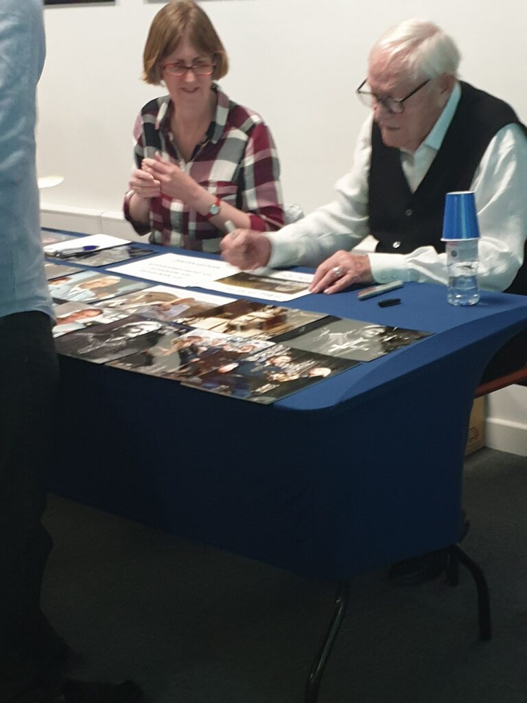 Julian Glover sitting at a table signing autographs