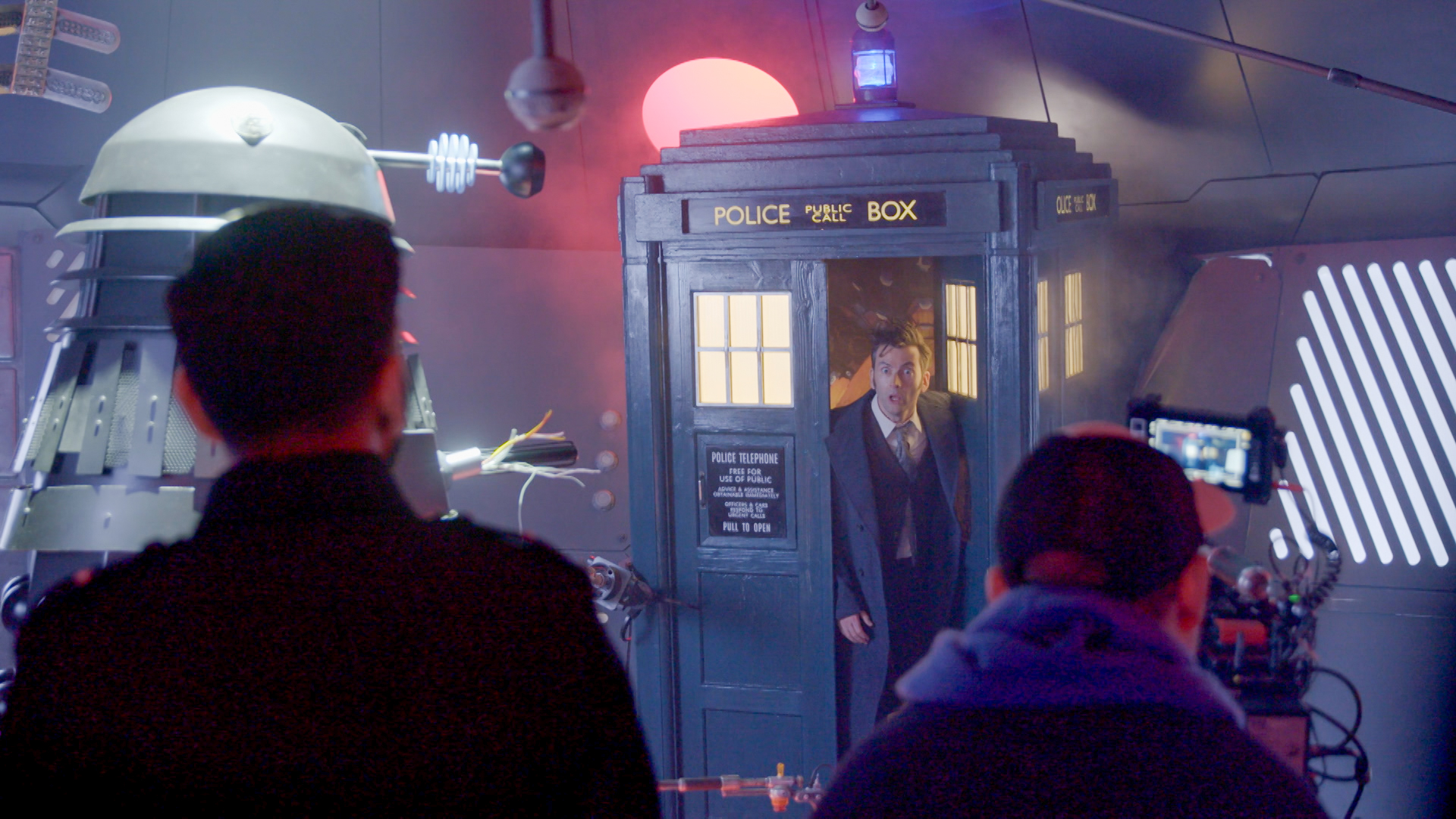 David Tennant on set with a TARDIS and Dalek prop, surrounded by film crew.
