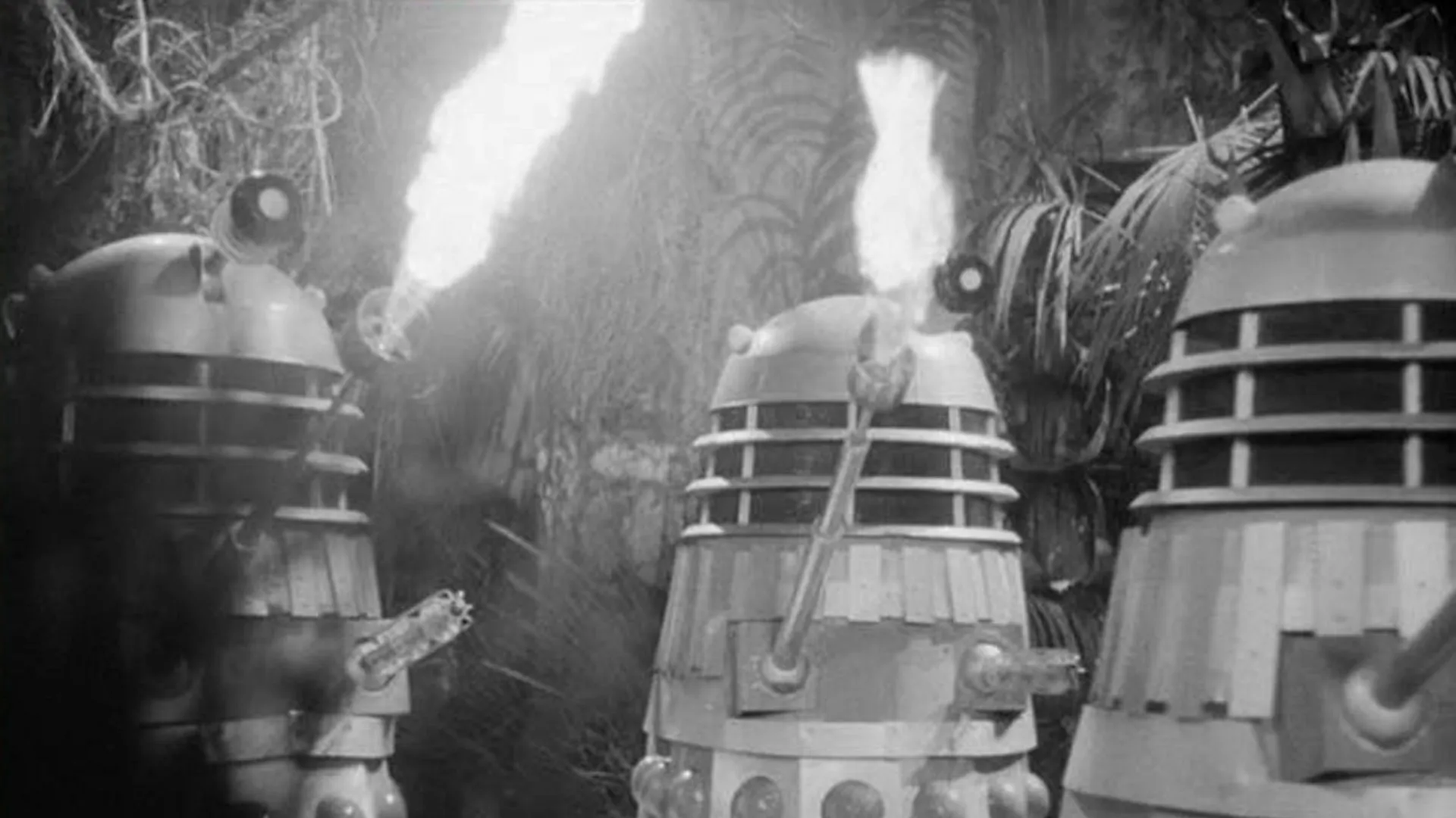 Three Daleks in a jungle; two of them are blowing fire out of their arm.